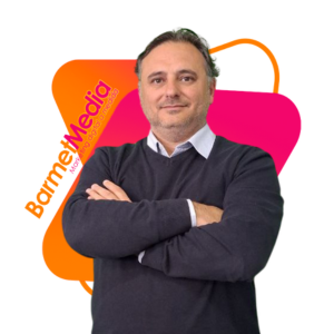 Rubén Salcedo con camisa blanca, jersey gris oscuro, delante de logotipo de BarmetMedia con diseño moderno de color naranja y rosa.